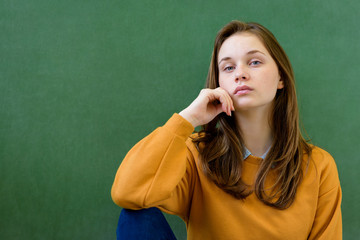 Young depressed female high school student sitting in front of chalkboard in classroom, with her hand on chin. Waist up portrait.