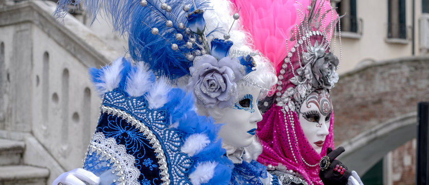 Two ladies holding fans, and wearing hand painted masks and ornate blue and pink costumes at Venice Carnival. Photographed in Campo Santa Maria Formosa.