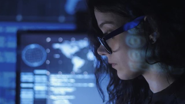 portrait of young woman programmer in eyeglasses working at a computer in the data center filled with display screens