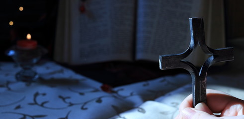 A young girl or a boy holds a cross in hands and prays at night. The background is an open old Bible book and nearby is candle with flame.
