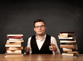 A young teacher in glasses sitting at classroom desk with pile of books in front of clean blackboard back to school concept.