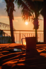 Still life. Smartphone and mug on bed amid evening sunset and silhouettes palms. Warm orange light. Concept nightfall. Copy space.