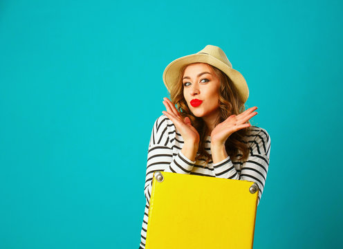 Portrait of young beautiful blond woman in hat with yellow suitcase over blue background