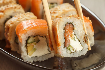Two chopsticks holding Uramaki with Conger and different  sushi rolls with seafood on ceramic plate on the background