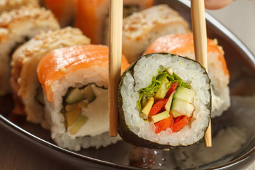 Two chopsticks holding Hosomaki with vegetables and different  sushi rolls with seafood on ceramic plate on the background