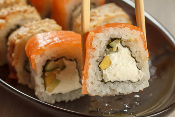 Two chopsticks holding Uramaki Philadelphia and different  sushi rolls with seafood on ceramic plate on the background