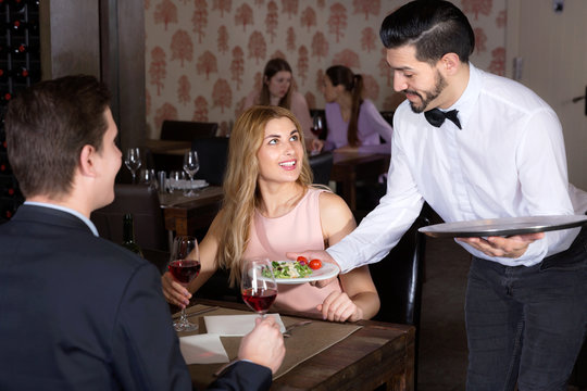 Polite waiter bringing ordered dishes to smiling couple at restaurant