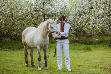 man dressed in national Ukrainian clothes holds a white horse in a flowering garden