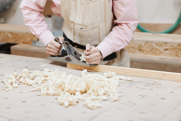 close-up of a male worker in a carpentry shop holds a plane in the hands. on a table of wood are...