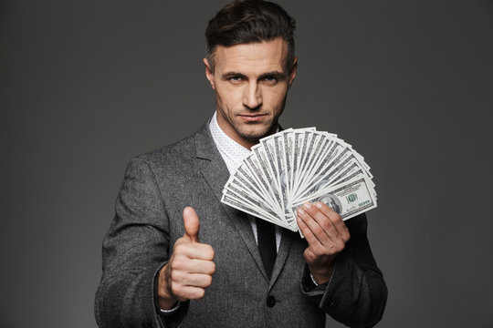 Photo of rich man 30s in business suit holding fan of cash money dollar currency and showing thumb up, isolated over gray background