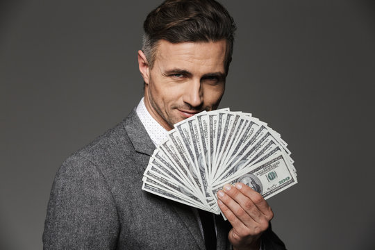 Photo of rich and happy entrepreneur guy 30s in business suit demonstrating money prize in dollar cash and looking on camera, isolated over gray background