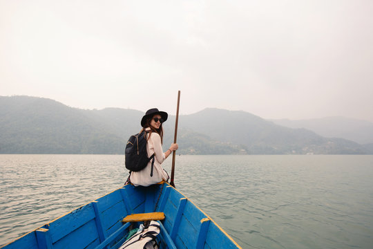 Woman wearing black hat and backpack sitting on boat with paddle, nice mountain lake Fewa and sunset on background