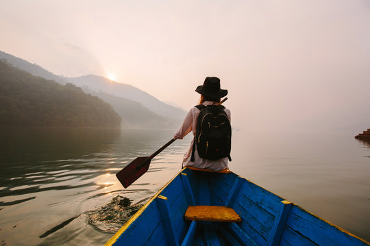 Woman wearing black hat and backpack sitting on boat with paddle, nice mountain lake Fewa and sunset on background