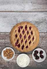Vertical image of a fruit pie surrounded by cherries, almonds and cream, taken on a wooden background 