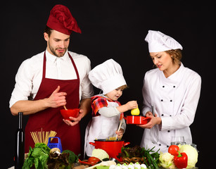 Adorable kid in chef hat with her parents. Mother and father teaching boy how to cook. Concept of friendly family, mutual assistance, support, cooking - happy family in kitchen. Healthy food at home.