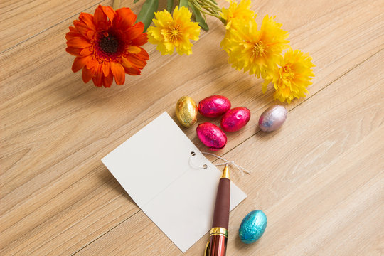 Chocolate Easter Eggs Over Wooden background with blessing card with pen and flowers on wood table.