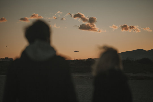 Man And Woman Silhouette While They Walk In Nature, With An Orange Sky And A Plane Flying In The Background.