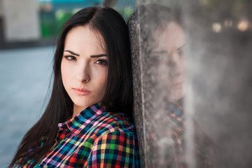 Fototapeta premium Portrait of young hipster teenager with long hair leaning against the wall in the street, wearing colorful shirt in cage on the urban background