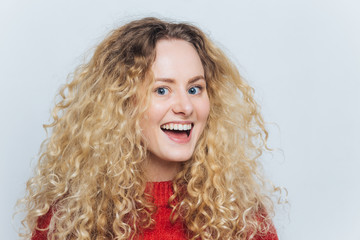 Headshot of beautiful satisfied female with pleasant joyful expression, with curly bushy hair, dressed in red sweater, doesn`t expect to hear something excited, isolated over white background