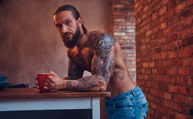 A handsome bearded tattoed male with a stylish haircut and muscular body, drinks coffee, leaning on a table.