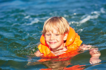Little boy in orange life vest swimming in wave sea