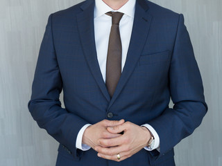 Torso of anonymous businessman standing with hands clenched in middle position wearing beautiful fashionable classic navy blue suit against grey backgound.