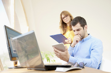Business team working at the office. Young financial businessman sitting in front of laptop and using digital tablet while businesswoman consulting with him. 