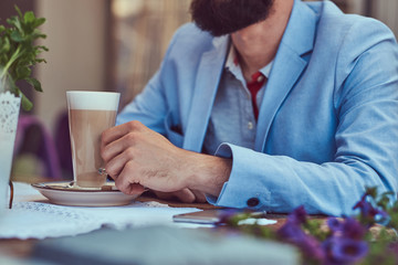 Portrait of a fashionable bearded male with a stylish haircut, holds a glass of a cappuccino, sitting in a cafe outdoors.
