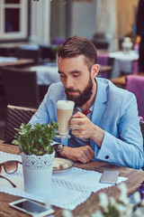 Portrait of a fashionable bearded male with a stylish haircut, drinks a glass of a cappuccino, sitting in a cafe outdoors.