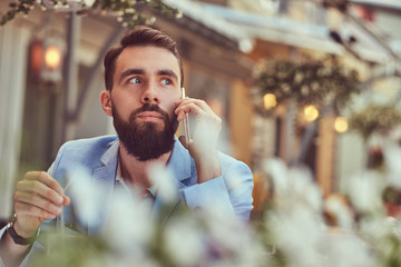 Close-up portrait of a fashionable bearded male with a stylish haircut, speaking by phone, sitting in a cafe outdoors.