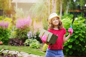 child girl playing little gardener and helping in summer garden, wearing hat and gloves, working with tools