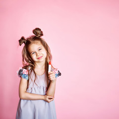 Happy children, little amazing girl with long brunette hair, looking at camera and smiling, holding a red lipstick in hand, isolated on a pink background.