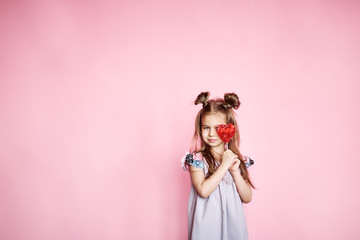 Cheerful little girl with long brunette hair in a dress holding a candy in the form of a heart in hands isolated on a pink background. Celebration of bright carnival for children, birthday