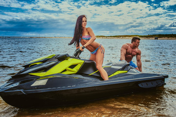 Attractive couple of a sexy girl and shirtless muscular male have fun with a jet ski on a seacoast.