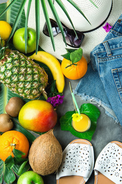 Flat Lay Arrangement Composition with Tropical Summer Fruits Glass of Fresh Juice Pineapple Mango Bananas Coconut on Large Palm Tree Leaf. Women Jeans Shorts Slippers Hat Sunglasses. Vacation