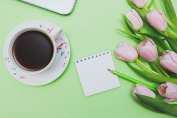 Female blogger workplace with laptop, pink tulips, tea cup, open notebook and green pencil on light green background. Top view with empty space for your text, greetings