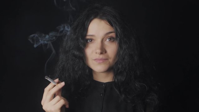 Portrait of young brunette woman with curly hair is smoking a cigarette and looking at the camera on the dark background