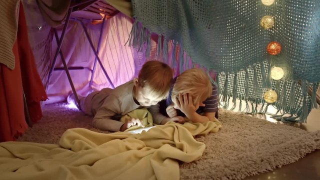 Two little boys lying on floor inside of cozy handmade play tent decorated with fairy lights and using digital tablet