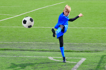 Teenage blonde Caucasian soccer (football) player in blue sport uniform is kicking ball in corner kick during game on grass field on sunny day