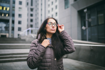 Portrait beautiful young business woman brunette in jacket and sweater stands on background office building, business center in glasses for view. Hand holding onto shackle of glasses and looking away