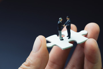 Business success strategy with collaboration, teamwork or negotiation jigsaw key, miniature people businessmen handshaking on white jigsaw puzzle piece in real human hand, dark black background