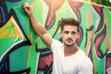 Fototapeta na wymiar Attractive muscle man leaning on colorful graffiti wall, wearing white t-shirt