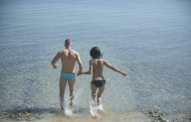 Couple in love running into sea water, sea surface on background, rear view. Couple topless run into sea with splashes of water. Vacation concept. Young family on honeymoon going to swim in ocean.