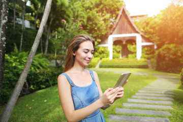 bright portrait of young woman using presses tablet and smiling on green palms and house background with sunshine. Concept of international travel to Thailand and new technologies