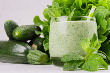 Green vegetable smoothie in glass with deep green vegetables and straw, mint, closeup. Soft white wooden board background.