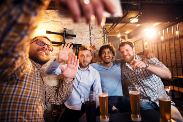 Four smiling multicultural friends waving and taking a selfie while sitting in the pub.