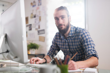 Looking at camera. Young hipster man sitting at his desk