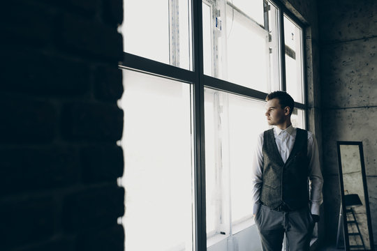 Young man dressed in a suit staying near a big window in a gray office and looking away .