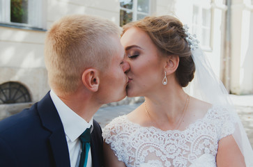 Beautiful blonde couple is sitting on wooden bench in old city. Groom in dark blue suit and bride in lace satin wedding dress talking in ancient town. Stone walls and pink rose bouquet. Smile and love