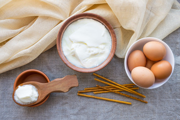 Rustic products: homemade fermented milk kefir (sour cream) in a wooden spoon and clay bowl, some brown eggs and biscuit sticks on linen cloth. Rustic natural organic food